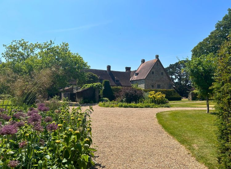 Gravel pathway winding through landscaped gardens with flowering borders, lawns, and a historic building in the background.